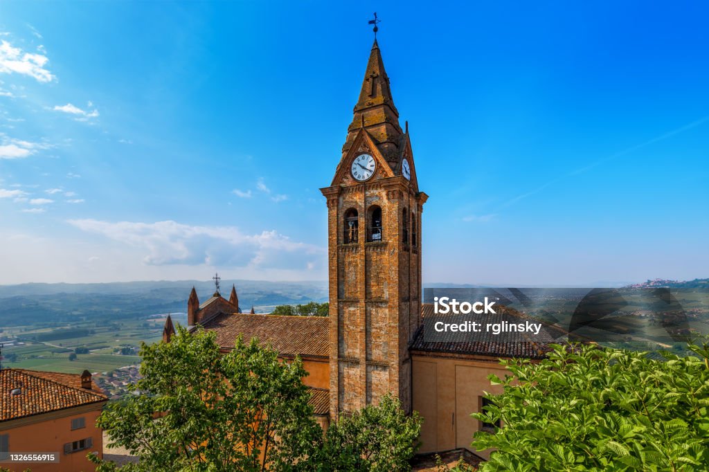 Old brick belfry under blue sky in Magliano Alfieri, Italy. View from above on church's roof and old brick belfry with clock under blue sky in small town of Magliano Alfieri in Piedmont, Northern Italy. Architecture Stock Photo
