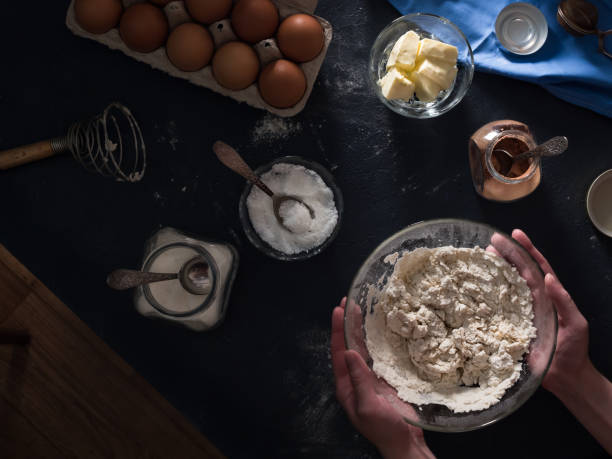 istock Cooking a cake. On the table are ingredients for making a pie (flour, eggs, cocoa, butter, sugar, salt), kitchen utensils and textiles. Kneading the dough. Hands in the frame. Top view of the table and floor. Flat lay. Dark background 1326575167