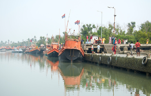 Fraserganj Fishing harbour, West Bengal, 01-24-2021: Traditional fishing trawlers docked along bank of Edward Creek. Also seen are few travellers standing near jetty, who come to visit the harbour and near fish market.
