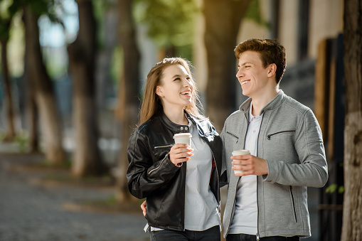 Young happy beautiful couple walking in the street. They are drinking coffee, hugging and laughing.