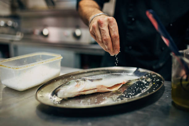 chef assaisonne le bar avec une pincée de sel - prepared fish photos et images de collection