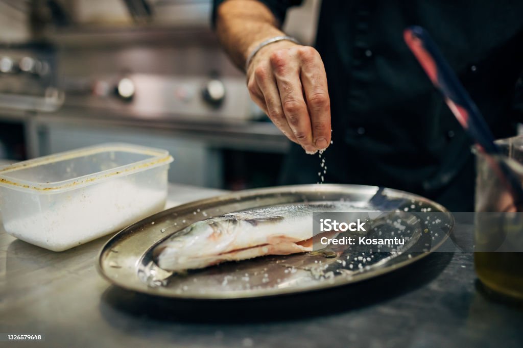 Chef seasoning the sea bass with a pinch of salt In the modern kitchen, a professional male chef, season the sea bass with salt, to gain an extra taste of the fish Fish Stock Photo