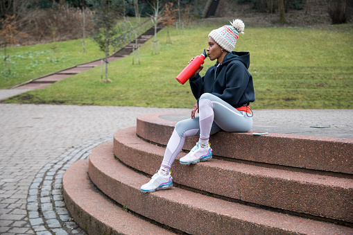 Black woman in sportswear sitting on a step in a local public park in her town. She is drinking from a sustainable flask.