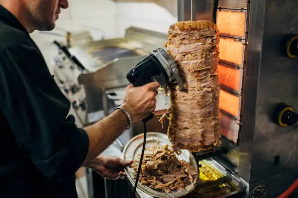 Professional male chef, in the kitchen, using the electric tool/rotated knife to cut the kebab/gyros, while meat spinning and grilling on the gyros grill