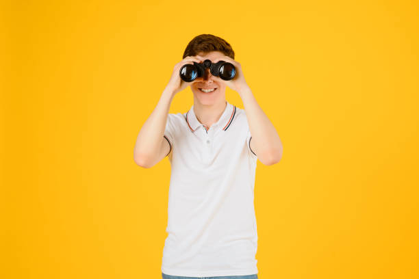 Portrait of a young happy man in white t-shirt smiling and looking through binoculars on a yellow background. Spyglass tourism concept. Portrait of a young happy man in white t-shirt smiling and looking through binoculars on a yellow background. Spyglass tourism concept. Copy space for your text binoculars point of view stock pictures, royalty-free photos & images