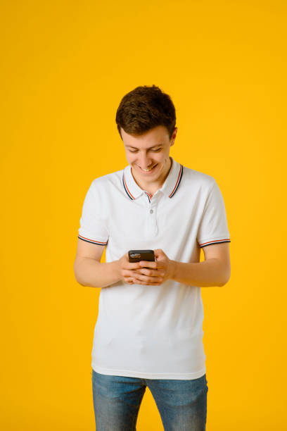 istock Young man in white t-shirt is using his smartphone and laughing on a yellow background. 1326606304
