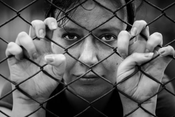 Suffering woman trapped behind wire fence Monochrome image depicting a woman imprisoned behind a wire fence. Her hands are grasping the chicken wire and the woman's sad, forlorn face is looking directly at the camera. people trafficking stock pictures, royalty-free photos & images