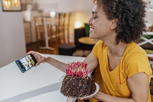 A Mixed race woman is in the living room, she is celebrating her birthday via video call on a mobile phone