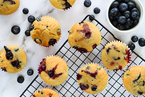 Image of black, metal cooling rack containing batch of freshly baked blueberry muffins in paper cake cases besides bowl of fresh blueberries, on marble effect background, elevated view