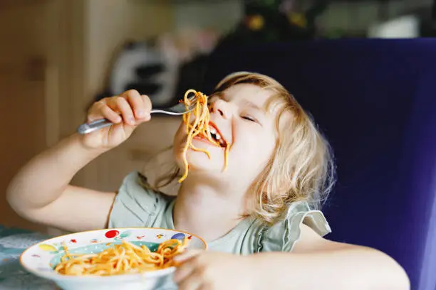 Photo of Adorable toddler girl eat pasta spaghetti with tomato bolognese with minced meat. Happy preschool child eating fresh cooked healthy meal with noodles and vegetables at home, indoors.