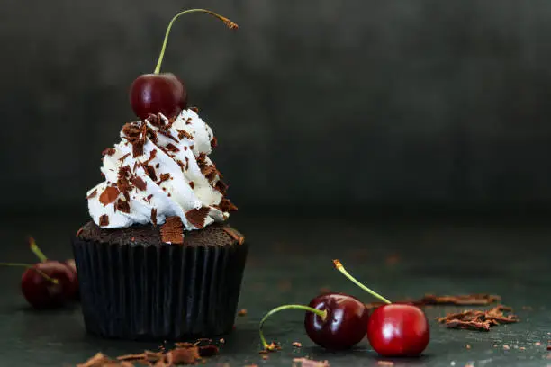 Photo showing an individual freshly baked, homemade, Black Forest gateau cupcake in brown paper cake cases against a black background. The cup cake has been decorated with piped whipped cream rosette topped with a morello cherry and sprinkled with chocolate shavings.