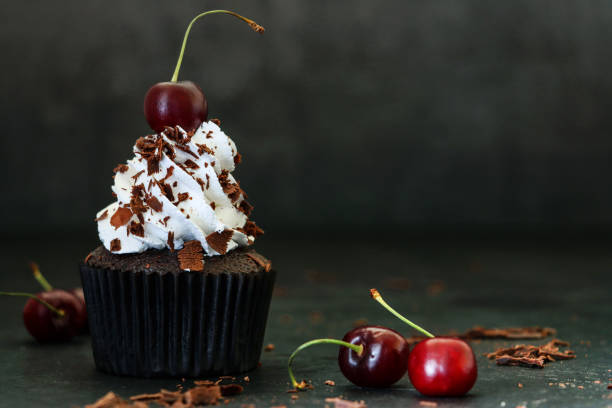 image of individual, homemade, black forest gateau cupcake in brown paper cake case, piped whipped cream rosette topped with morello cherry sprinkled with chocolate shavings, black background, focus on foreground - cake chocolate cake chocolate gateaux imagens e fotografias de stock