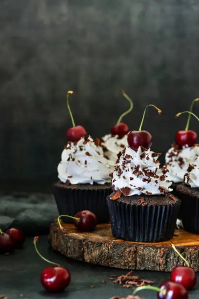 Photo showing a wooden cake stand of freshly baked, homemade, Black Forest gateau cupcakes in brown paper cake cases against a black background. The cup cakes have been decorated with piped whipped cream rosettes topped with morello cherries and sprinkled with chocolate shavings.