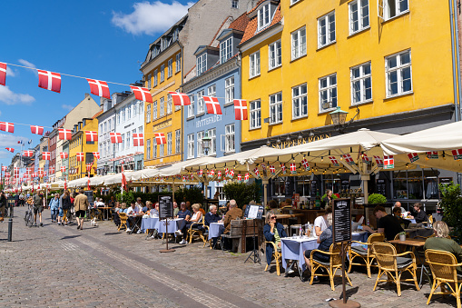 Copenhagen, Denmark - 13 June, 2021: people enjoy a summer day in the busy Nyhavn quarter on the waterfront in Copenhagen with many restaurants and bars and Danish flags above