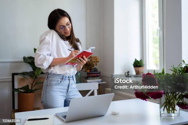 Cheerful Young Beautiful Woman Talking On The Phone And Using Laptop Stock Photo - Download Image Now
