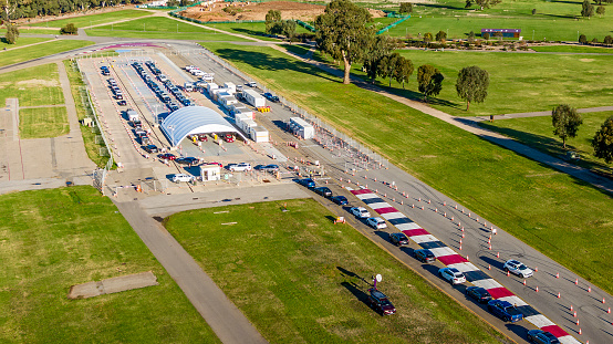 Aerial view pop-up Covid-19 testing centre & cars in parklands on the edge of the city of Adelaide, South Australia: large canopy set on former racetrack with rows of people in cars, SUVs, taxi, waiting to receive a Covid-19 test.  Logos and ID edited.