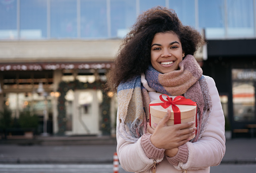 Beautiful African American woman holding gift box, standing on urban street and smiling. Portrait of young emotional female looking at camera with birthday present in hand