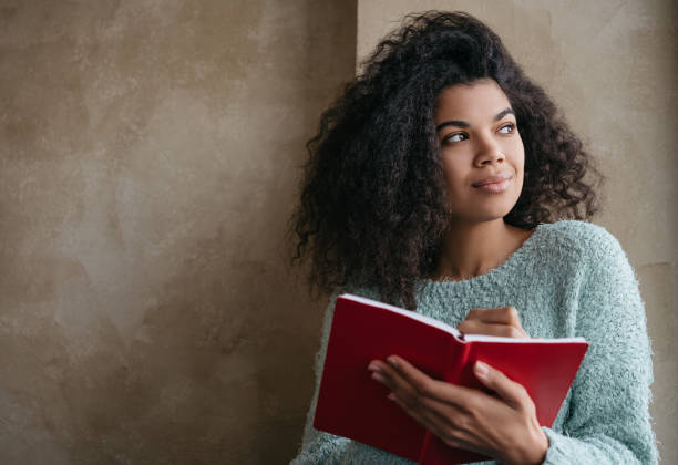 bella donna afroamericana che tiene in mano un libro rosso, guardando la finestra e sorridendo. studente universitario che studia, impara la lingua, siede in biblioteca. ritratto di giovane scrittore pensieroso che prende appunti - creativity thinking author writing foto e immagini stock