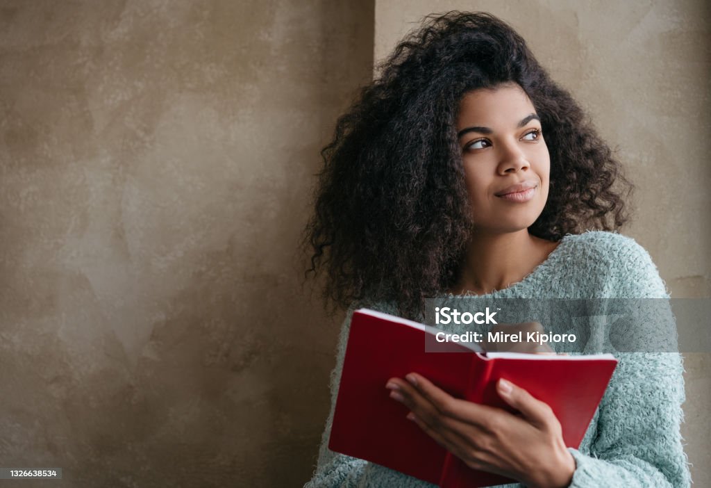 Beautiful African American woman holding red book, looking at window and smiling. University student studying, learning language, sitting at library. Portrait of young pensive writer taking notes Young beautiful African American woman holding red book, looking at window and smiling. University student studying, learning language, sitting at library. Portrait of young pensive writer taking notes Writing - Activity Stock Photo
