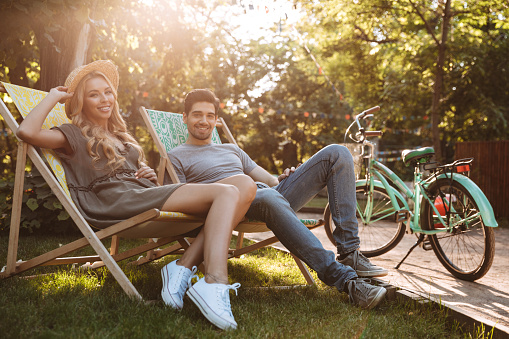 Side view of Happy lovely young couple sitting together on sun loungers and looking at the camera outdoors