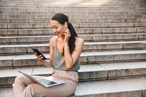 Image of beautiful smiling woman 20s wearing casual summer outfit and bluetooth earphone sitting on street stairs and holding smartphone while using silver laptop
