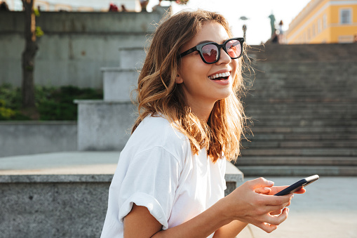 Picture of european brunette woman wearing casual summer outfit and sunglasses laughing while walking through city street with smartphone in hands