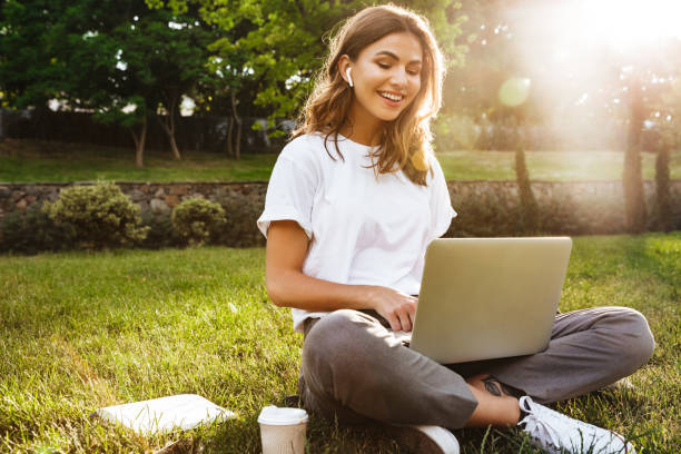 여름철에는 다리가 교차한 공원의 녹색 잔디에 앉아 있는 예쁜 젊은 여성의 초상화가 노트북과 무선 이어폰을 사용하여 화상 통화를 했습니다. - laptop computer grass nature 뉴스 사진 이미지