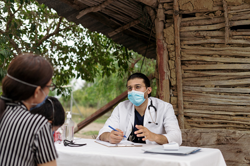 A latin male doctor wearing a face masks and talking to patients in a rural area for consultation.