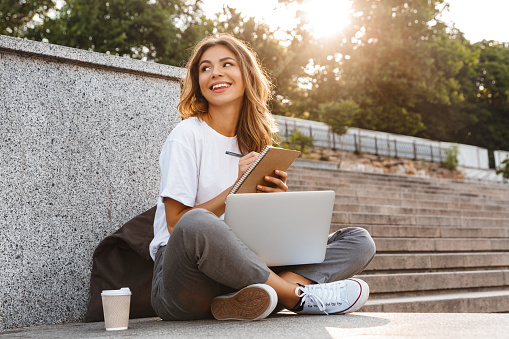 Smiling young girl making notes in notepad while sitting outdoors with laptop computer and coffee cup