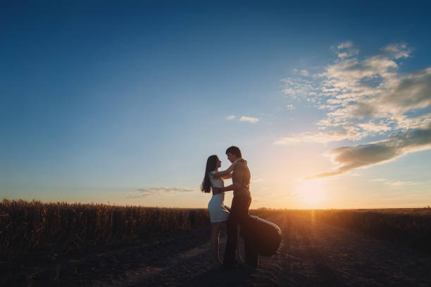 deux jeunes amoureux mariés se tiennent debout et s’embrassent avant un baiser sur fond de coucher de soleil. silhouette d’un gars avec une guitare et d’une fille sur la route entre les champs de blé. style champêtre. far west. - back to front photos et images de collection