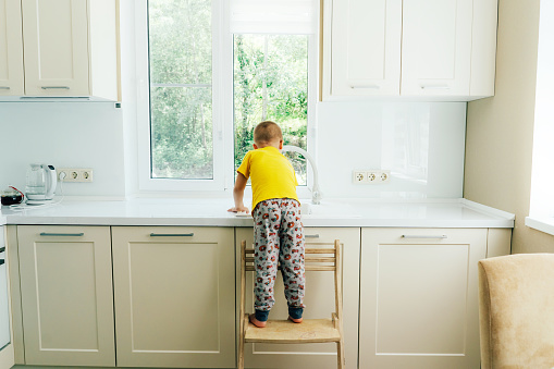 Little boy standing on a chair washes the dishes in the sink in the kitchen, back view.