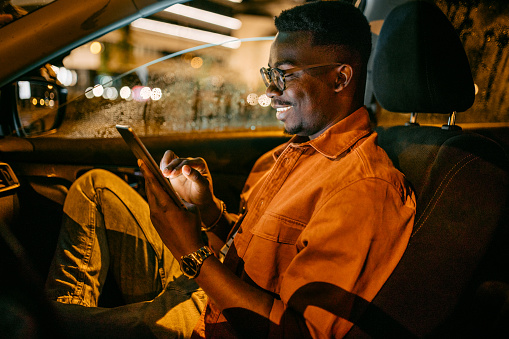 Young handsome black businessman working in car on his digital tablet during journey in rainy night.