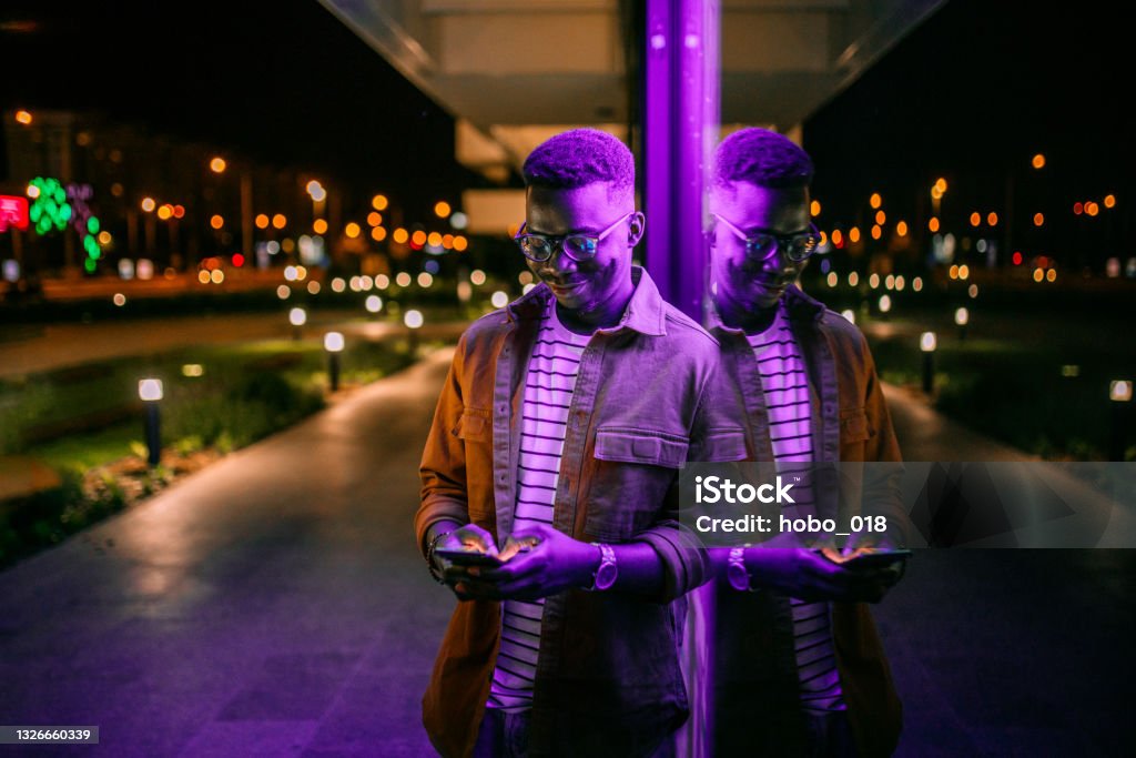 Using Phone in a front of neon lights on the street Young handsome black man using phone, typing text message in a front of neon blue light office window in rainy night People Stock Photo