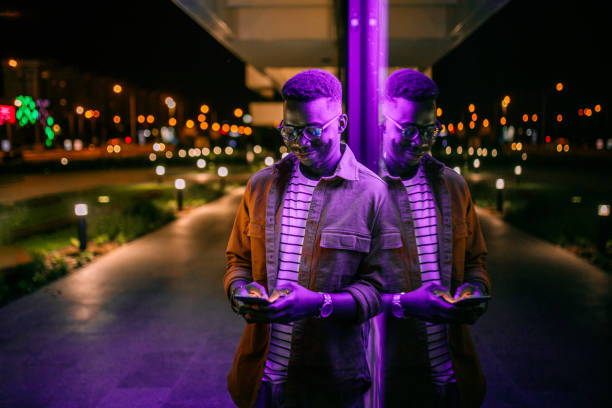 Using Phone in a front of neon lights on the street Young handsome black man using phone, typing text message in a front of neon blue light office window in rainy night computer technology people stock pictures, royalty-free photos & images