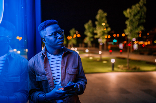 Young handsome black man using phone, typing text message in a front of neon blue light office window in rainy night