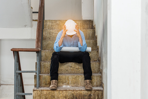 Asian construction engineer having problems at work sitting on stairway in construction site and holding head with hopelessly suffering emotional pain. Business fail concept.