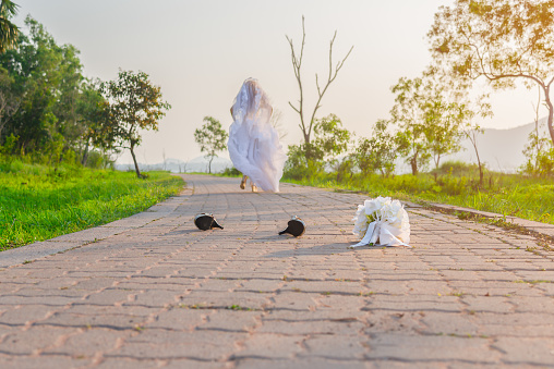 Beautiful bride wearing a white wedding dress running away alone in nature outdoor with leaving a bouquet of flowers and shoes on the street. Runaway bride before the wedding ceremony concept.