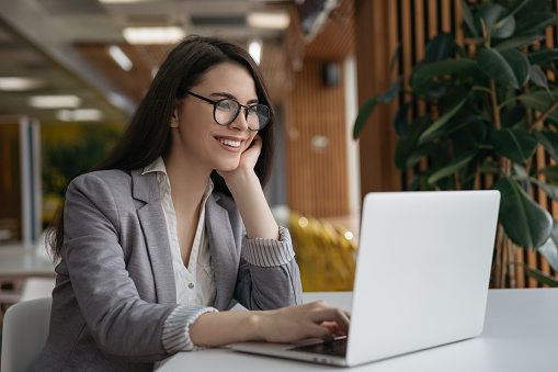 Smiling businesswoman using laptop computer, watching training courses. Portrait of happy successful manager working in office. Beautiful woman freelancer wearing stylish eyeglasses sitting at workplace