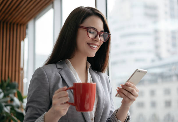 Young smiling businesswoman holding cellphone and cup of coffee, communication, checking email in office. Beautiful woman in stylish red eyeglasses using mobile app for online shopping. Freelancer receive payment empresaria sonriente sosteniendo celular y taza de café, comunicación, revisando el correo electrónico en la oficina. hermosa mujer en elegantes anteojos rojos utilizando la aplicación móvil para las compras en línea. freelancer recibe pago