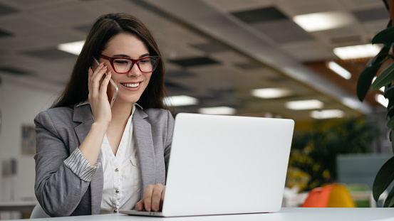Successful businesswoman talking on mobile phone, working in office. Woman freelancer in stylish eyeglasses using laptop computer, looking at digital screen, communication at workplace