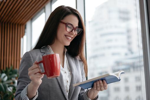 Beautiful woman in stylish eyeglasses reading book, holding cup of coffee, smiling. Portrait of young successful businesswoman planning start up project in office. Freelancer working from home