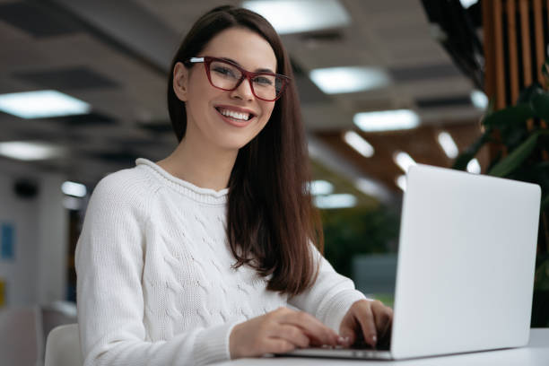 Happy copywriter working freelance project, typing, looking at camera and smiling. Attractive business woman using laptop computer, sitting in office. Student learning language online