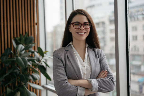 Young confident business woman with arms crossed standing in office. Successful manager in formal clothes and stylish eyeglasses looking at camera and smiling stock photo