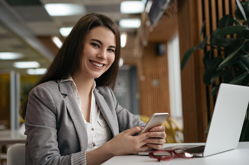 Beautiful smiling business woman using laptop computer, holding mobile phone working in office