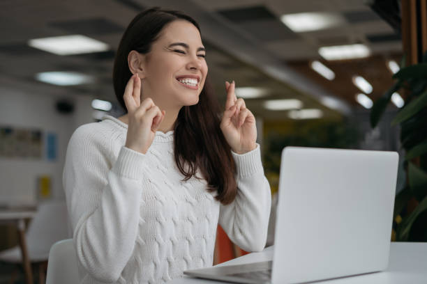 joven hermosa mujer sosteniendo los dedos cruzados para la buena suerte. estudiante emocional usando la computadora portátil y los resultados del examen en espera - fingers crossed fotografías e imágenes de stock