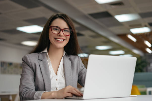 istock Young insurance agent using laptop computer, working at workplace. Successful business woman looking at camera and smiling, sitting in modern office 1326677417