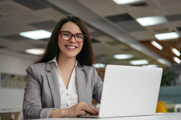 Young insurance agent using laptop computer, working at workplace. Successful business woman looking at camera and smiling, sitting in modern office stock photo