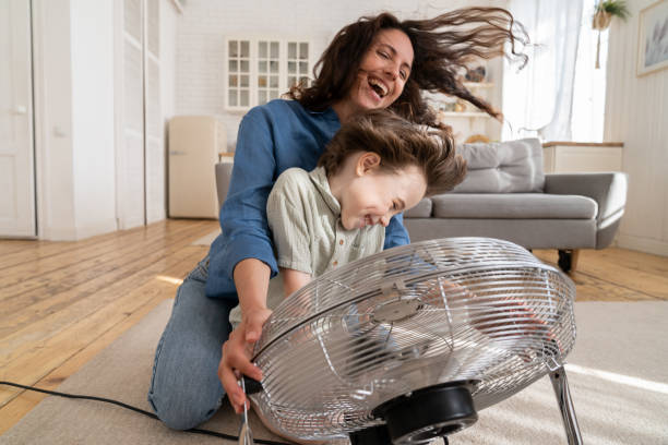 Joyful single mother bonding with son excited together at home with fan blowing wind in living room Playful single mother bonding with son excited playing together at home with big fan blowing cool wind in living room. Mum and child enjoy time together at conditioning ventilator during summer heat air conditioner family stock pictures, royalty-free photos & images