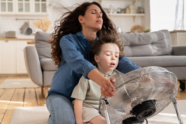 Relaxed mom and small kid refreshing sit at big indoor ventilator blowing cooling fresh air at home Happy mom and small kid refreshing sit near big indoor ventilator blowing cooling fresh air at home on floor bonding. Young mother with preschool kid enjoying conditioner having fun indoors together air conditioner family stock pictures, royalty-free photos & images