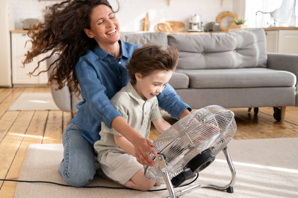 Caring mom relax has fun with child at home sit laugh together with son in front of fan ventilator Loving mom relax and having fun with child at home: carefree young woman mother sit laughing together with son in front of fan ventilator work indoors and blow cool air during summer heat outdoors air conditioner family stock pictures, royalty-free photos & images